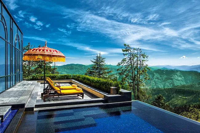 Poolside loungers under an umbrella overlook lush green mountains and a bright blue sky at a luxury resort.