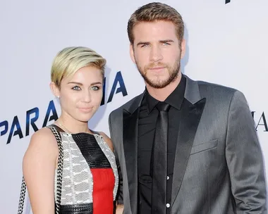 A woman with short blonde hair and a colorful dress stands beside a man in a gray suit and black tie at an event backdrop.