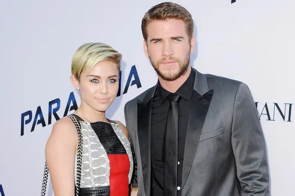 A woman with short blonde hair and a colorful dress stands beside a man in a gray suit and black tie at an event backdrop.