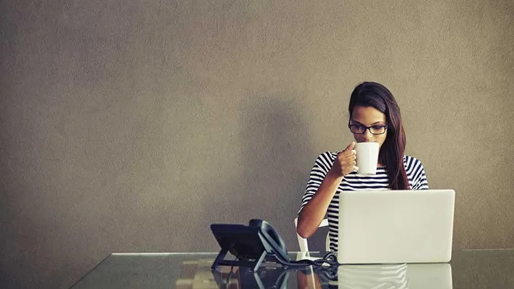 A young woman drinks coffee at her desk.