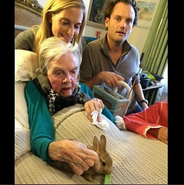 Four people gathered around an elderly woman in bed, holding a rabbit. The scene is warm and caring.