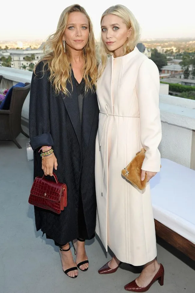 Two women in stylish long coats on a terrace, one in black holding a red bag, the other in white holding a tan clutch.
