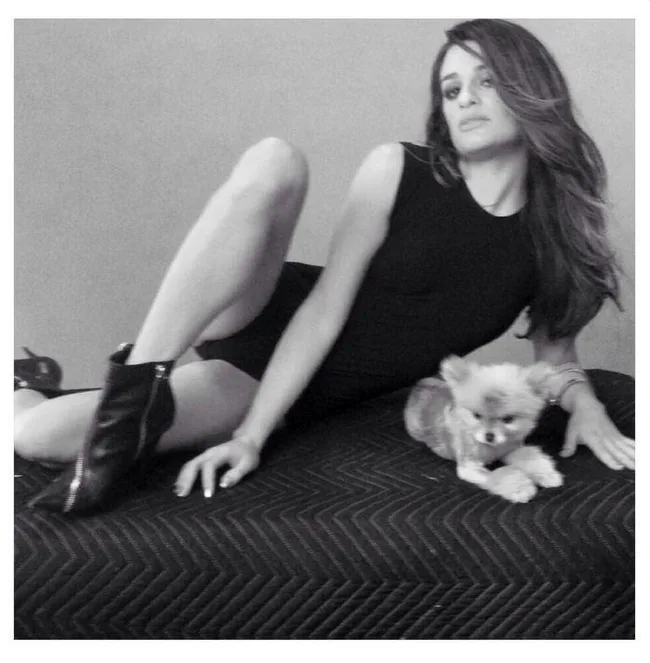 A woman in a black dress poses on a quilted surface with a small fluffy dog next to her. Black and white photo.
