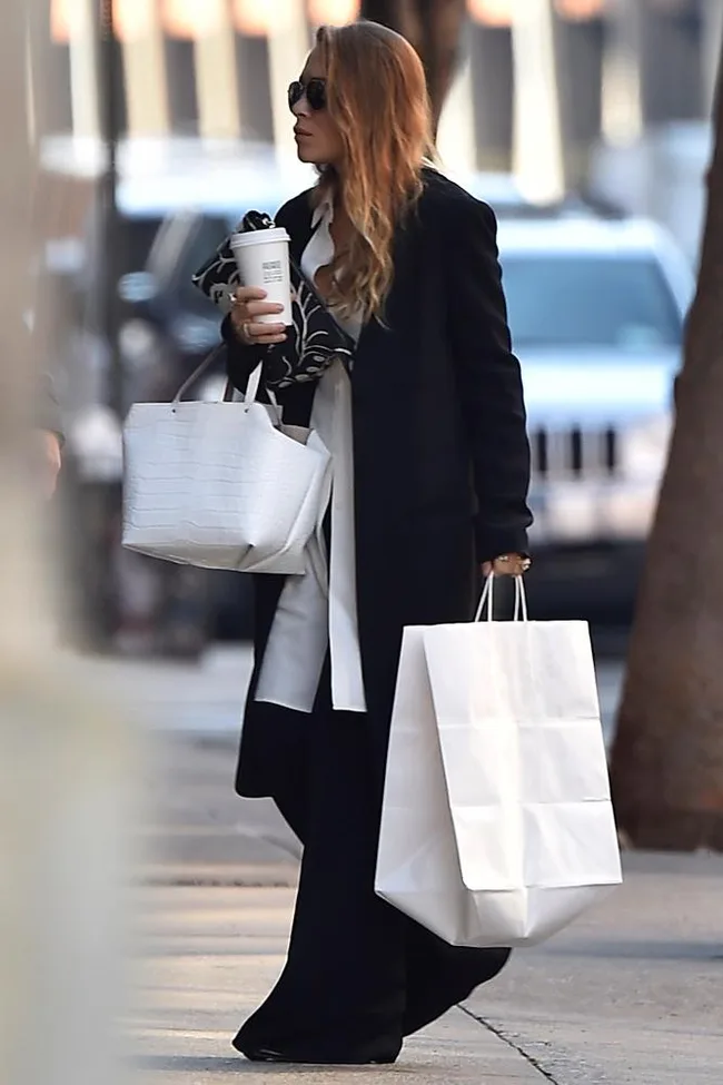 A woman in a black coat carries a large white shopping bag and a coffee cup, walking on a city street.