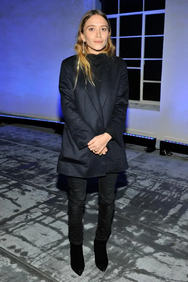 Young woman in a dark coat and boots stands indoors on a concrete floor with blue lighting in the background.