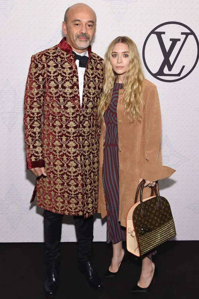 Two people pose at a Louis Vuitton event, one in a red patterned coat, the other in a brown coat with a designer bag.