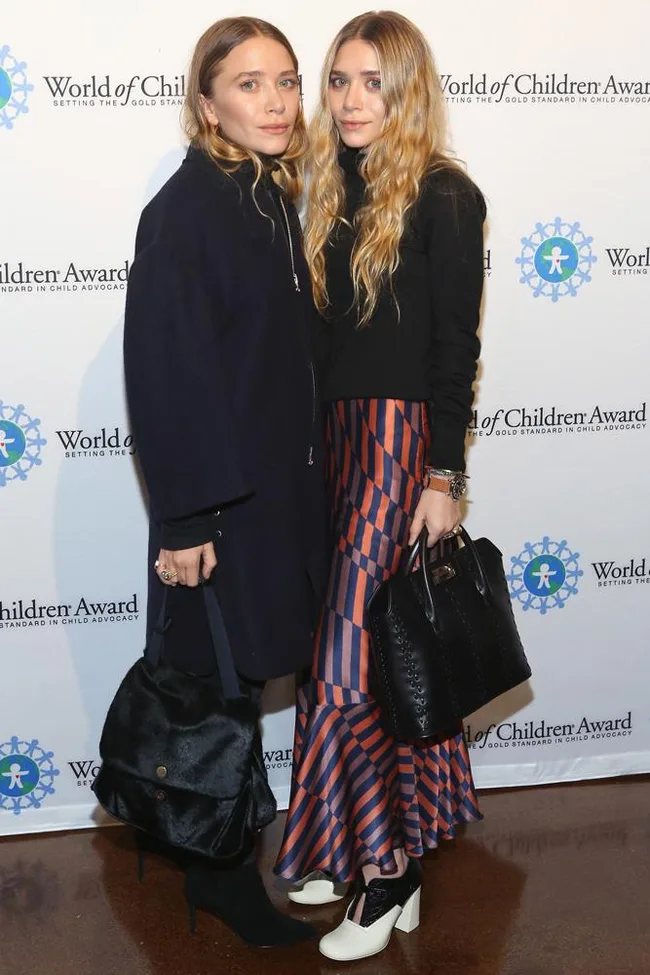 Two women pose together at the World of Children Awards, wearing stylish outfits and holding black handbags.