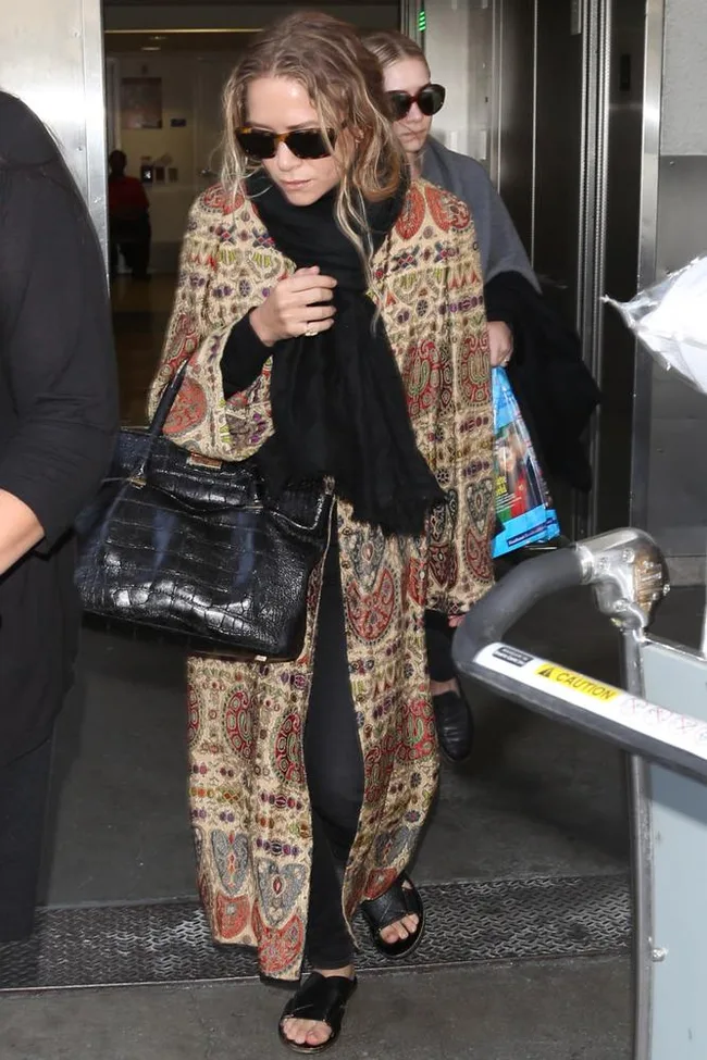 Woman in sunglasses, wearing a patterned coat and black accessories, walks through an airport with another person.