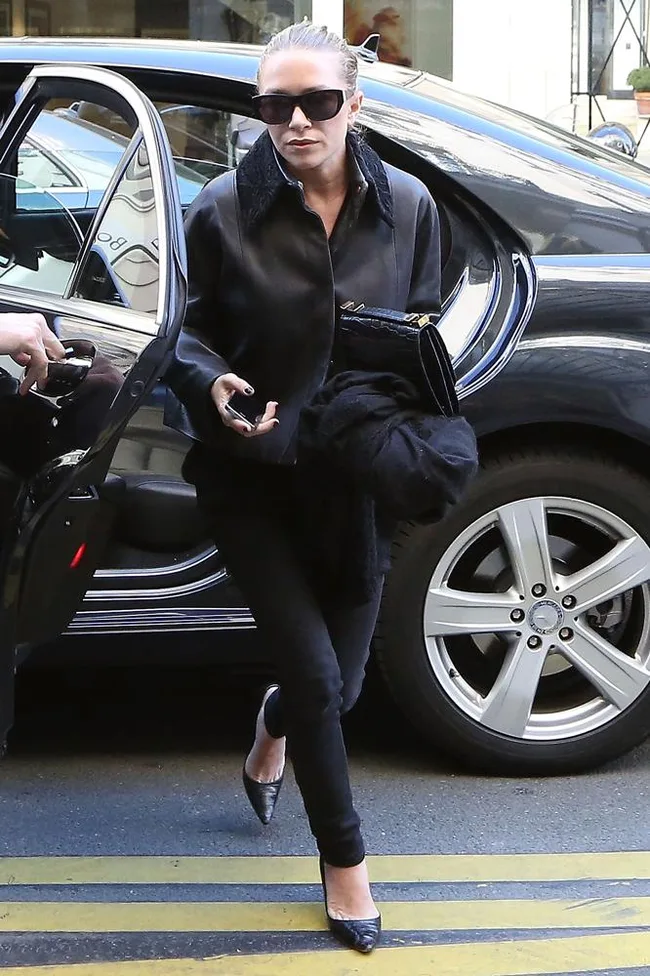 ```plaintext
Person in dark outfit and sunglasses exits car, holding clutch, on a city street.
```