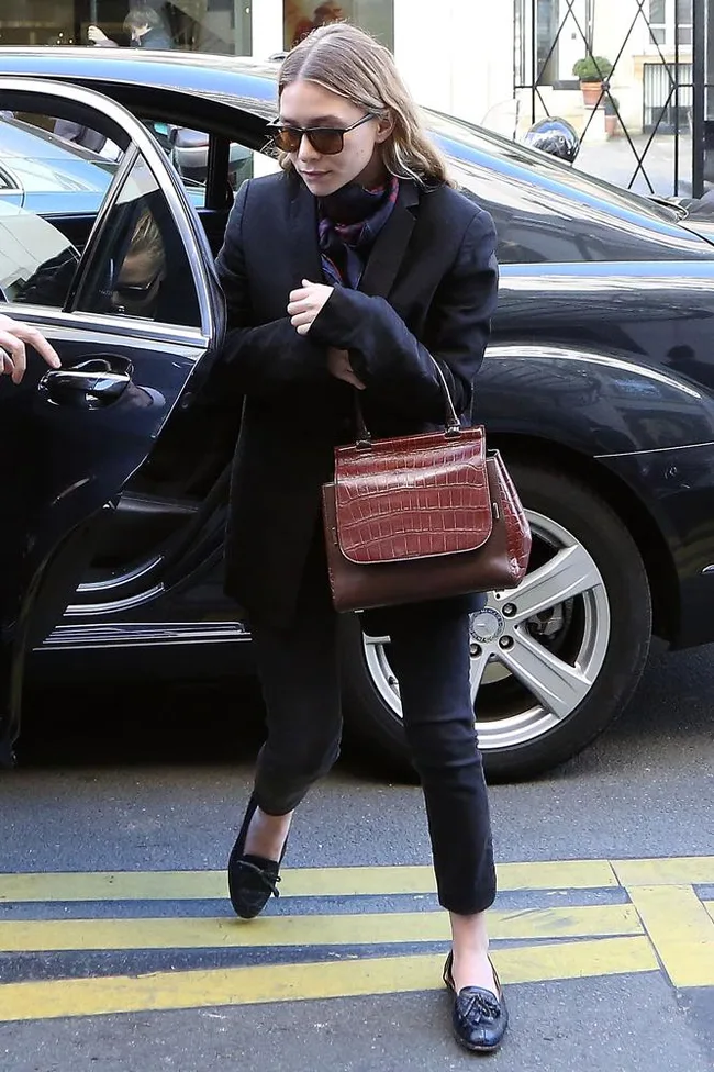 Person in black blazer and sunglasses exits car, holding a brown handbag.