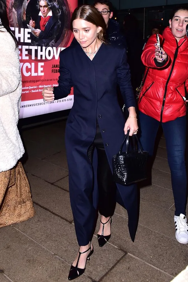 Woman in a dark coat and heels walking with a black handbag, surrounded by people in a public setting.