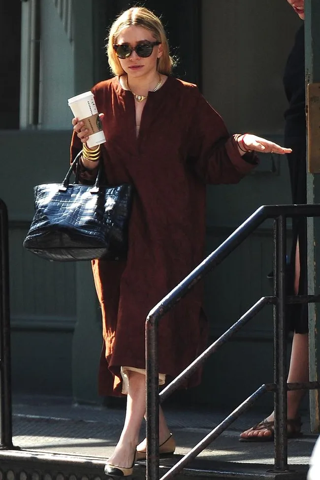 A woman in sunglasses and a brown dress holds a drink and a large handbag while walking down steps.