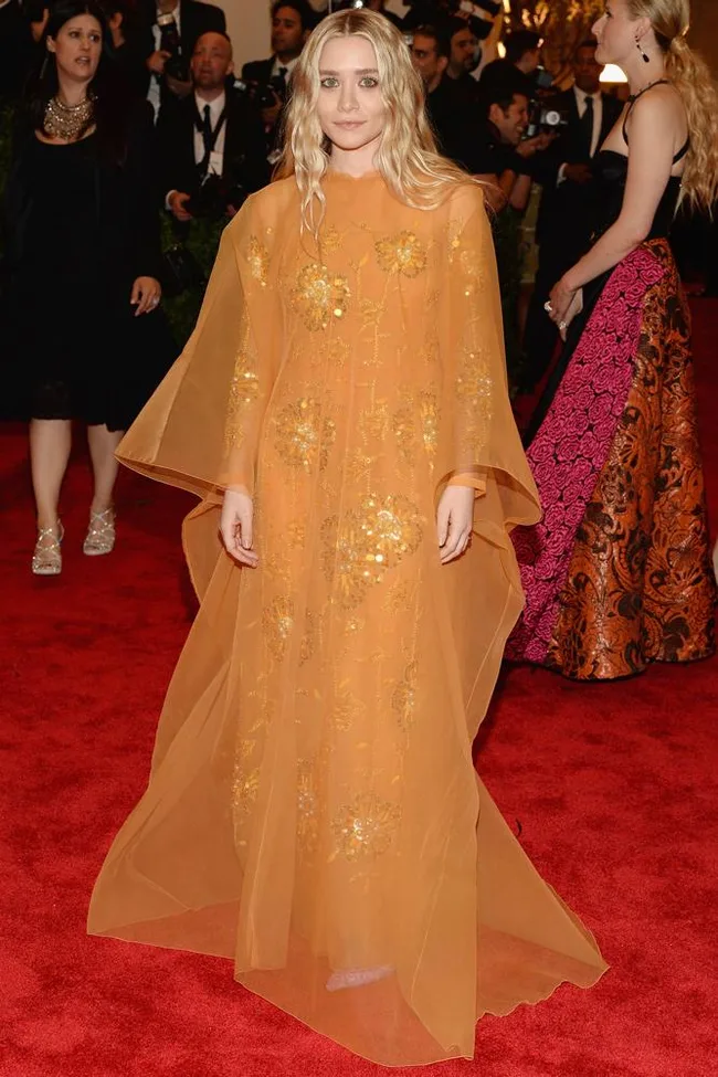Person in an orange floral gown on a red carpet with crowd in the background.
