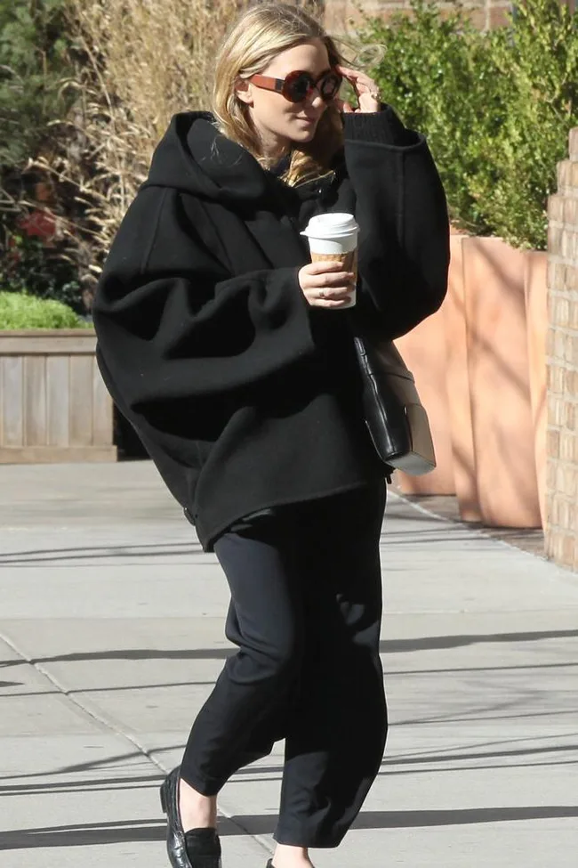 Person in oversized black coat, sunglasses, holding a coffee cup, walking on a sunny street.
