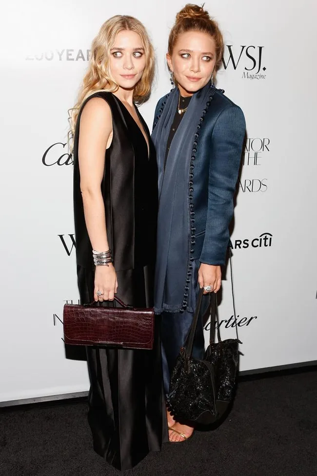 Two women on a red carpet, one in a black dress and the other in a blue outfit, both holding handbags.