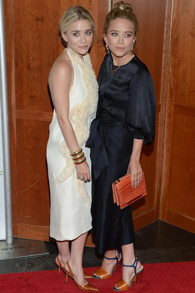 Two women on a red carpet, one in a white dress and the other in a black dress holding an orange clutch.