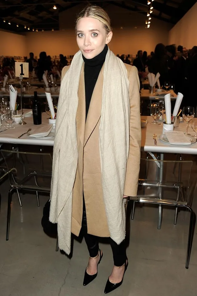A person in a tan coat and scarf stands indoors near set dining tables, with people conversing in the background.