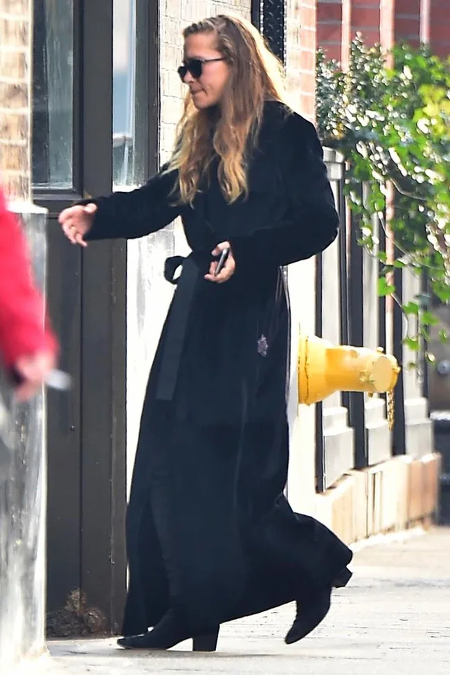 A woman in a black coat and sunglasses walks on a sidewalk near a yellow fire hydrant.
