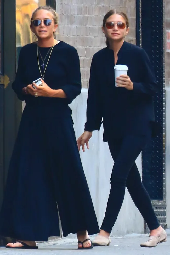 Two women in sunglasses, wearing black outfits, walking on the street. One holds a phone, the other a coffee cup.