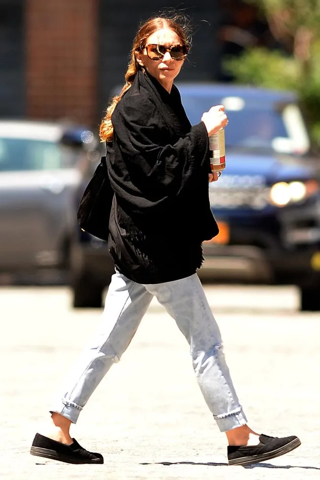 A person walking outdoors wearing sunglasses, a black shawl, light jeans, and holding a can.