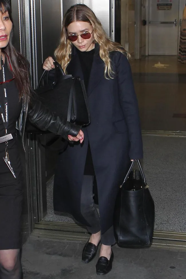 A woman in a dark coat and sunglasses carries two large black bags, walking through a door.