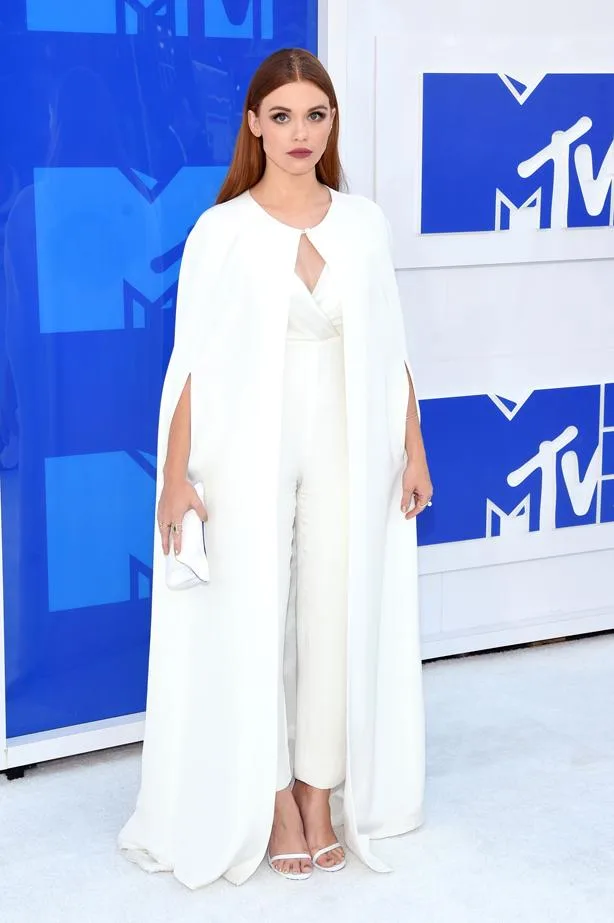 A woman in a white outfit and cape stands on a white carpet at an MTV event.