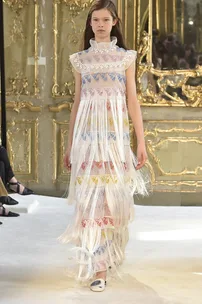 Model in a sheer, tiered fringe dress with colorful embroidery patterns, walking on a runway with ornate gold backdrop.