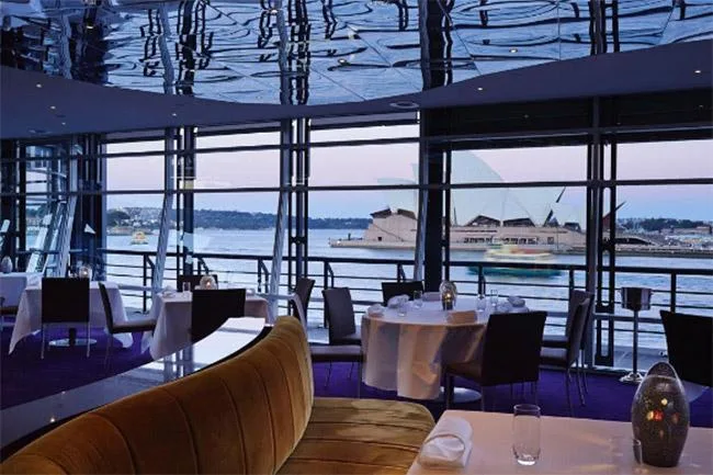 Inside a modern restaurant with a view of the Sydney Opera House across the water through large windows.