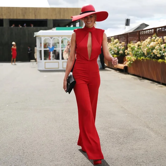 Woman in a red jumpsuit and wide-brimmed hat walks outdoors smiling, holding a black clutch purse.