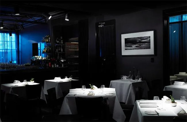 Dimly lit elegant restaurant interior with white tablecloths, a bar in the background, and blue lighting accents.