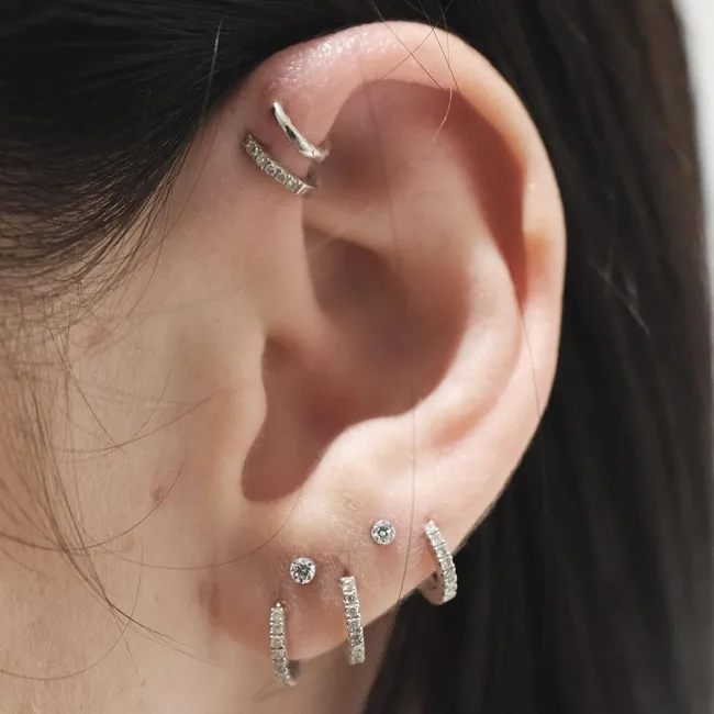 A close-up of an ear with multiple piercings, featuring silver hoop earrings and small stud earrings.