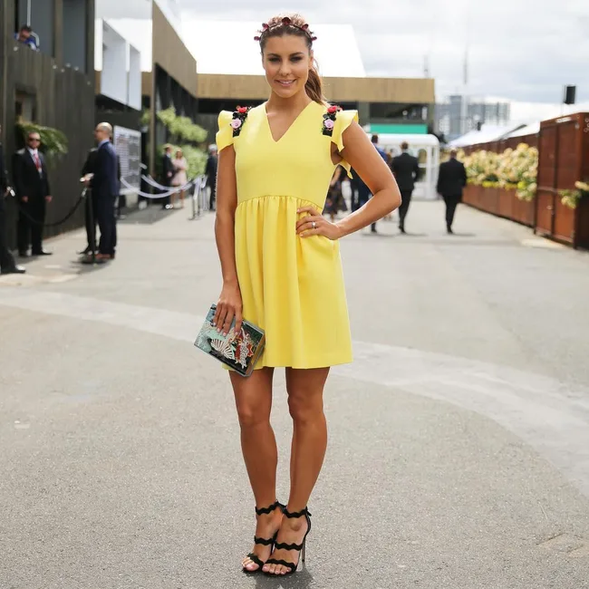 A woman in a yellow dress poses outdoors, holding a clutch, with a floral headpiece and black sandals.