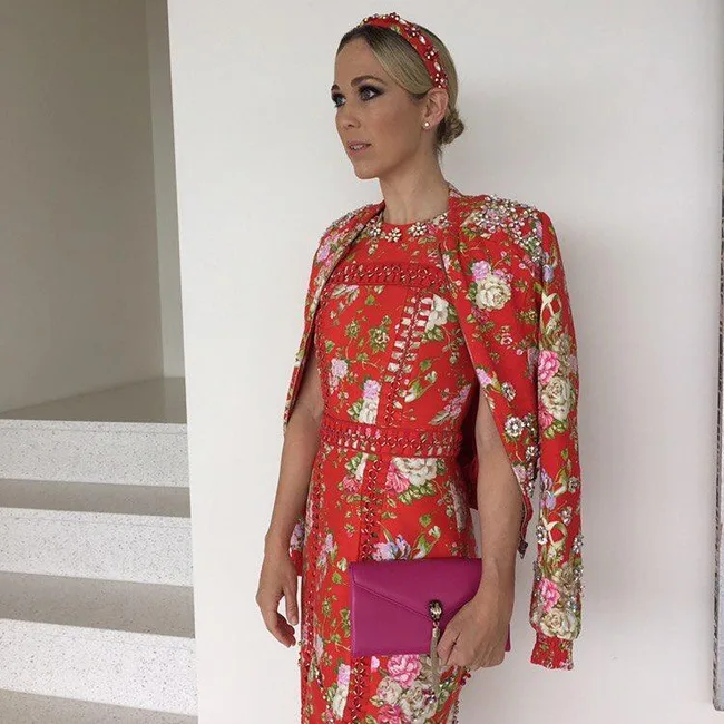 Woman in floral red dress with pink clutch, standing indoors.