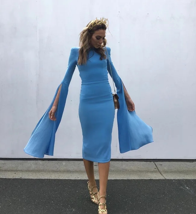 Woman in blue dress with cape sleeves and gold crown walks on a street.