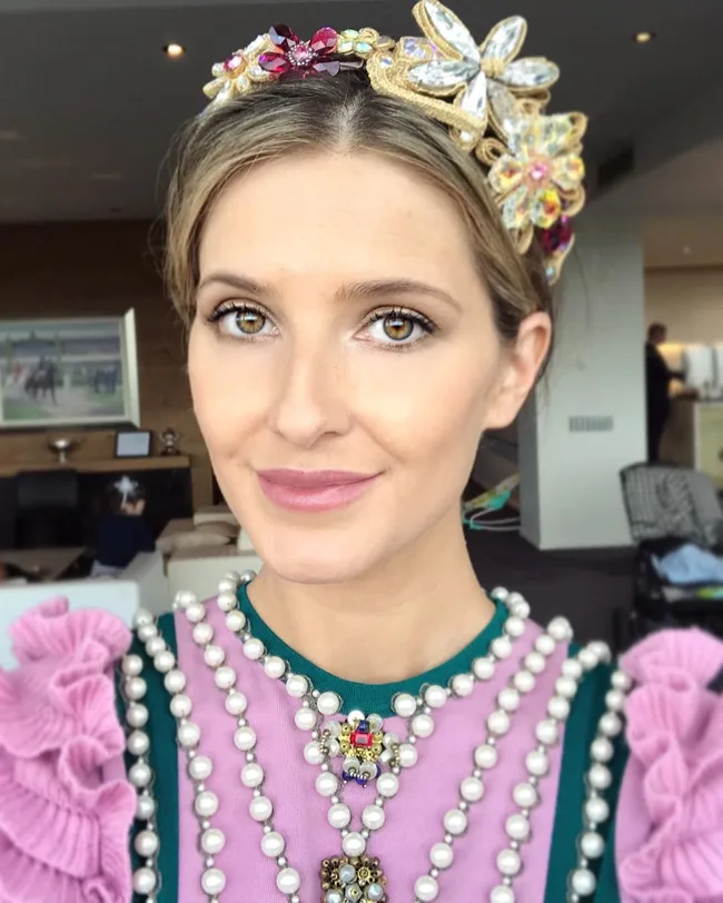 A woman with a jeweled headpiece and pearl necklace, wearing a pink top with ruffles, smiles slightly at the camera.