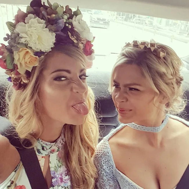 Two women with floral headpieces smile playfully in a car, one sticking out her tongue.