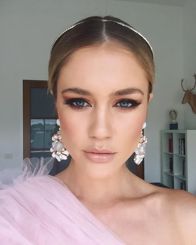 A woman with smoky eye makeup and pearl earrings wearing a pink dress with a tiara in a room with white walls.