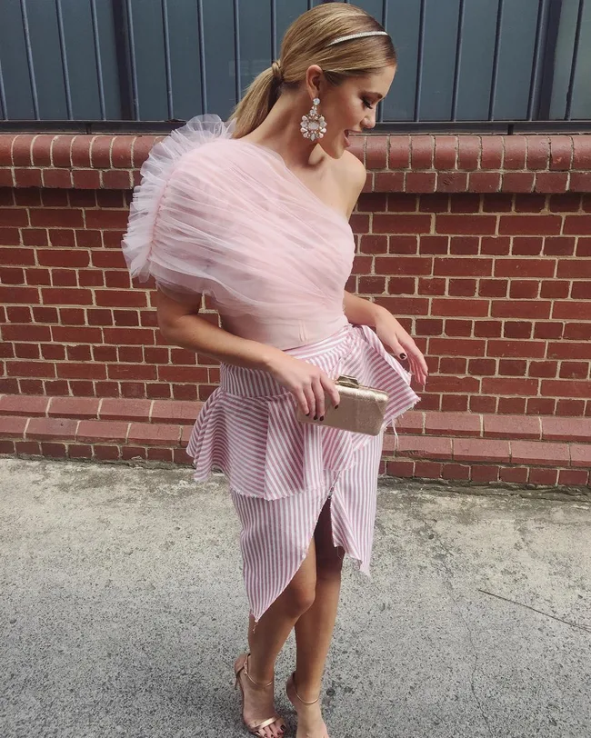 A woman in a pink striped one-shoulder dress holds a gold clutch against a brick wall.