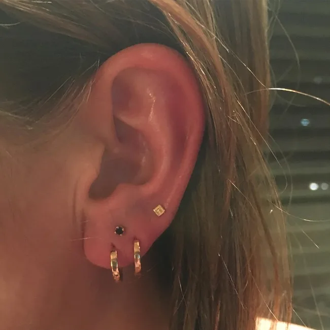 Close-up of a person's ear with multiple piercings, including gold hoops and small studs against a blurred background.