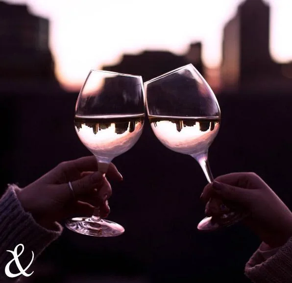 Two hands clinking wine glasses with a city skyline reflected, during a sunset.