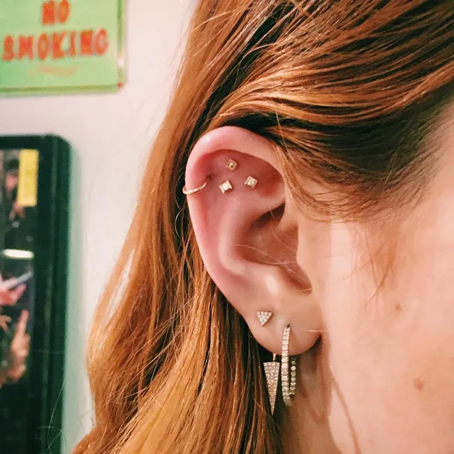 Close-up of a person's ear with multiple gold and diamond-shaped piercings; background shows a "No Smoking" sign.