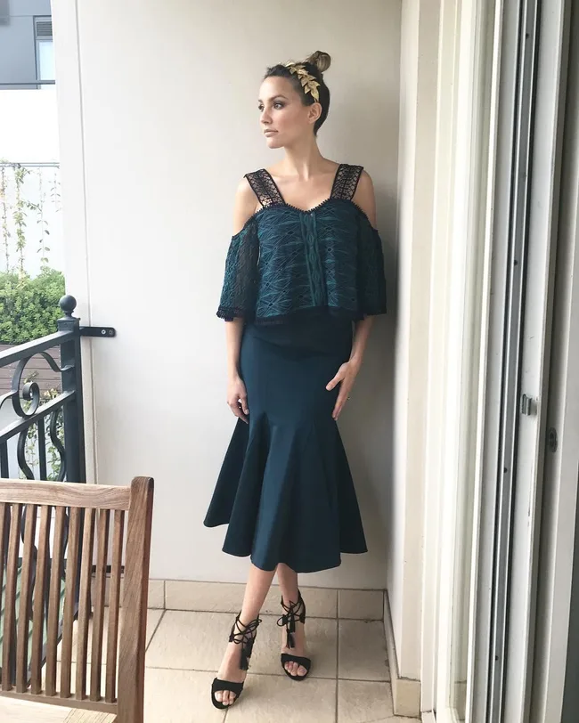 A woman in an elegant, off-shoulder dark teal dress with black heels stands on a balcony, looking to the side.