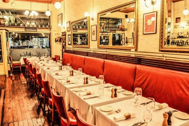 Cozy restaurant interior with red chairs, wooden floor, and a long table set for dining under warm lighting.