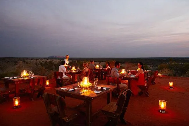 Outdoor dining setup on a desert landscape with people seated at wooden tables, illuminated by soft candlelight.