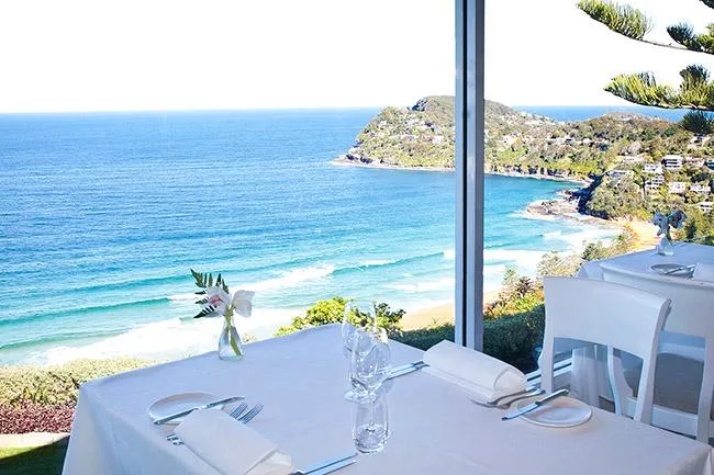 Restaurant table with ocean and coastline view through large windows. White tablecloths and set for dining.