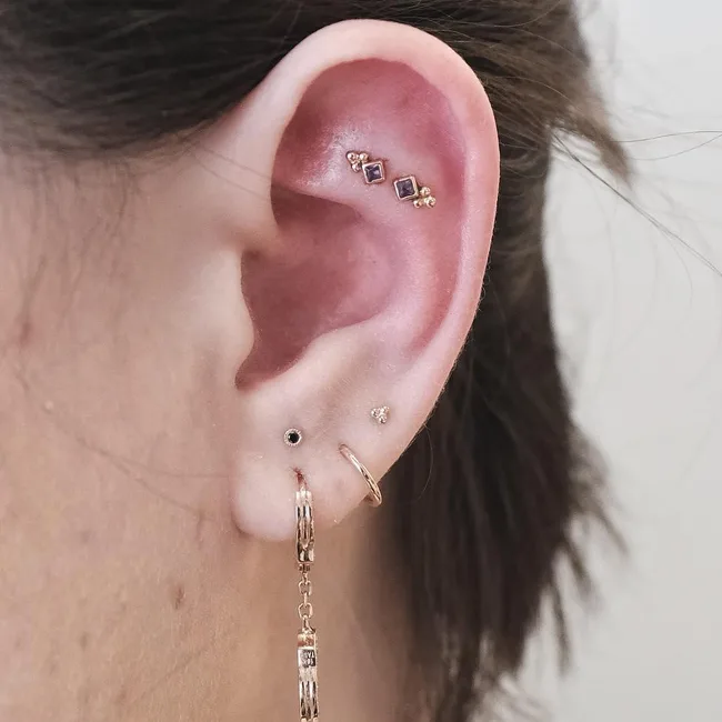 A close-up of an ear with multiple piercings and various earrings, including studs and a hoop with a chain.