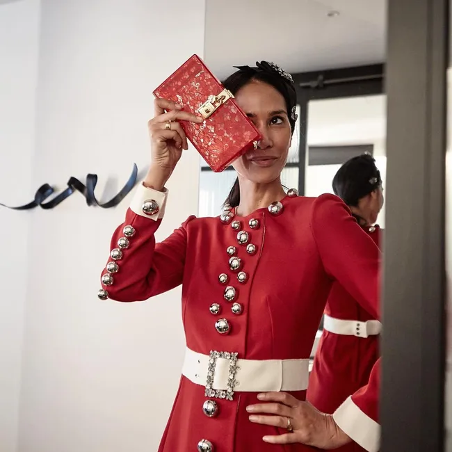 A woman in a red dress with silver buttons playfully holds a red clutch over her eye in a mirror reflection.