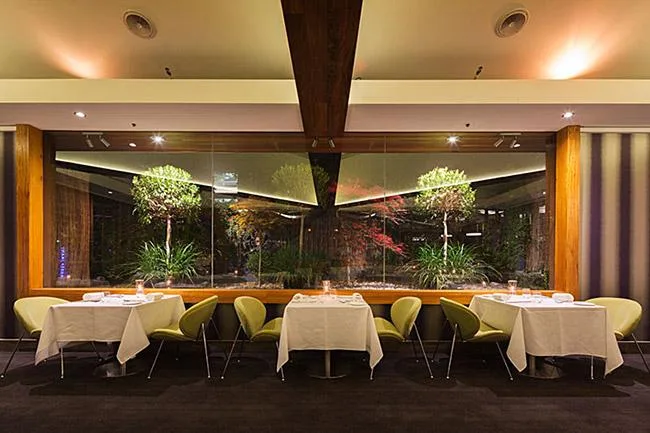 Elegant restaurant interior with green chairs, white tablecloths, and large window showcasing a lush garden view.