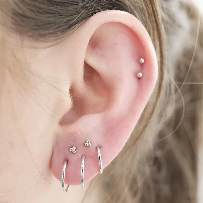 Ear with multiple piercings; features small stud and hoop earrings in silver.