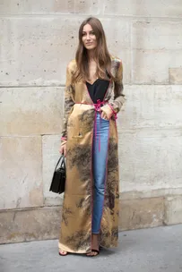 Woman in long, patterned silk robe over jeans and top, standing by a stone wall, holding a black handbag.